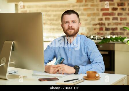 Ein netter junger Mann mit einem Bart in einem blauen Hemd, der mit seiner Kreditkarte vor dem Computer zu Hause posiert. Ein Kerl, der eine Online-Zahlung auf der Inter Stockfoto