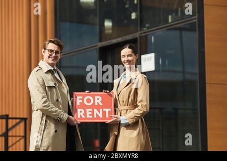 Porträt von zwei Immobilienmaklern halten rot FÜR MIETE Schild und lächeln an Kamera, während im Freien gegen moderne Bürogebäude, Kopierraum stehen Stockfoto