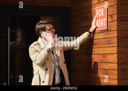 Taille-up-Porträt von männlichen Vermietmakler hängen FÜR MIETE Schild auf Bürogebäude während der Rede mit dem Smartphone im Freien, kopieren Raum Stockfoto