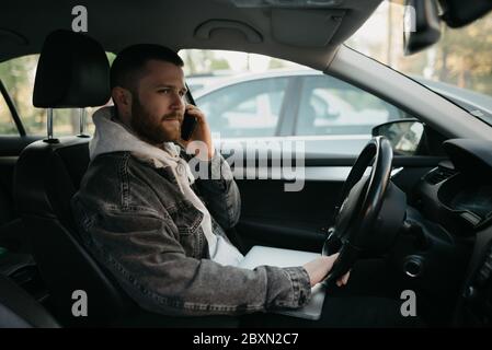 Ein Mann mit einem Bart in legerer Kleidung, der geschäftliche Anrufe auf seinem Smartphone macht, liegt in einem Auto ein Laptop auf seinem Schoß. Ein Kerl hielt sein Auto an Stockfoto