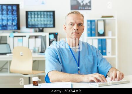 Porträt von schönen reifen Arzt Blick auf die Kamera, während am Arbeitsplatz sitzen und tippen mit Gehirn CT-Scans im Hintergrund, kopieren Raum Stockfoto