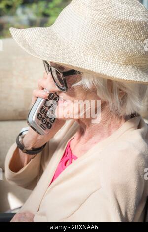 An ihrem Geburtstag, tragen Sonnenbrille und einen Sommerhut, sitzen draußen in der Sonne in ihrem Garten, genießen einen Fernruf von ihrer Enkelin. Stockfoto