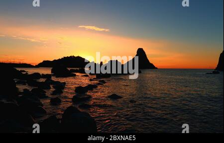 Faraglioni di Acitrezza - Silhouette con sagoma all'alba in controluce della scogliera con suggestive luci con riflesso sul mare e cielo blu Stockfoto
