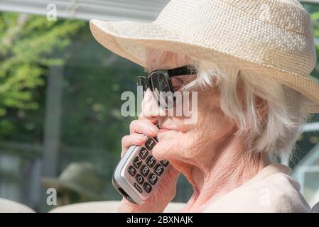 An ihrem Geburtstag, tragen Sonnenbrille und einen Sommerhut, sitzen draußen in der Sonne in ihrem Garten, genießen einen Fernruf von ihrer Enkelin. Stockfoto