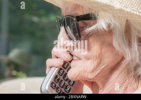 An ihrem Geburtstag, tragen Sonnenbrille und einen Sommerhut, sitzen draußen in der Sonne in ihrem Garten, genießen einen Fernruf von ihrer Enkelin. Stockfoto