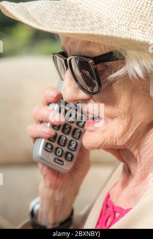 An ihrem Geburtstag, tragen Sonnenbrille und einen Sommerhut, sitzen draußen in der Sonne in ihrem Garten, genießen einen Fernruf von ihrer Enkelin. Stockfoto