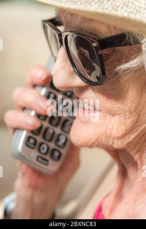 An ihrem Geburtstag, tragen Sonnenbrille und einen Sommerhut, sitzen draußen in der Sonne in ihrem Garten, genießen einen Fernruf von ihrer Enkelin. Stockfoto