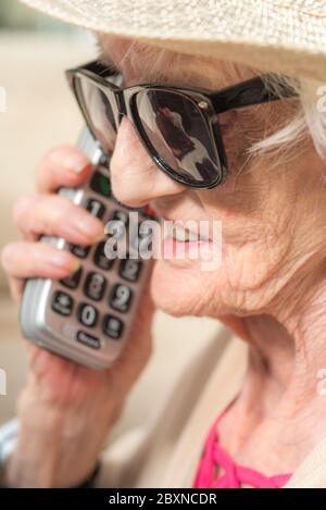 An ihrem Geburtstag, tragen Sonnenbrille und einen Sommerhut, sitzen draußen in der Sonne in ihrem Garten, genießen einen Fernruf von ihrer Enkelin. Stockfoto