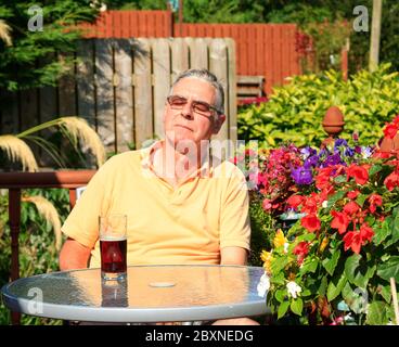 Ein zufriedener Mann mit Sonnenbrille saugt die Sonne auf, während er vor einem Glas Bier an einem Tisch sitzt, umgeben von Blumen Stockfoto
