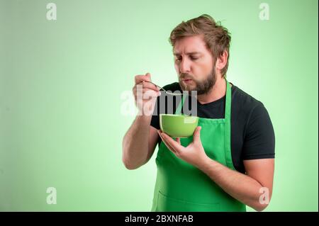 Supermarktmitarbeiter mit grüner Schürze und schwarzem T-Shirt, kühlen die Suppe, halten die Schüssel in der Hand isoliert auf grünem Hintergrund Stockfoto