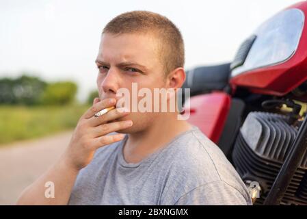 Ein junger Mann, der in der Nähe seines Motorrads sitzt und draußen raucht Stockfoto
