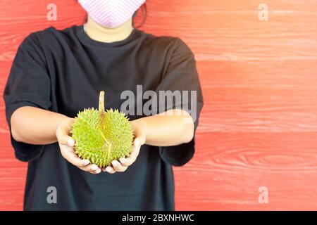 Junge Frau trägt eine Schutzmaske, um eine Infektion des Corona-Virus (Covid-19) in einem schwarzen Hemd mit einem reifen Durian zu verhindern, bereit, mit dem roten w zu essen Stockfoto