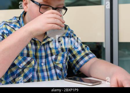 Rothaariger Junge mit Handy, der aus einem Glas Eiswasser mit Zitrone trinkt. Kind mit Brille in einem karierten Hemd gekleidet sieht auf das Smartphone und d Stockfoto