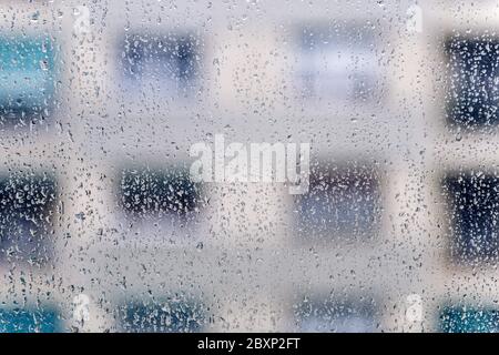 Tropfen Regenwasser, die durch das Glas eines Fensters fallen, im Hintergrund sind die Fenster des gegenüberliegenden Gebäudes außer Fokus Stockfoto