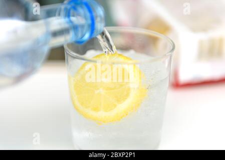 Mineralwasser mit Zitrone in das Glas gießen. Plastikflasche mit Mineralwasser, das mit Zitroneneis und Zitrone in das Glas gegossen wird. Stockfoto