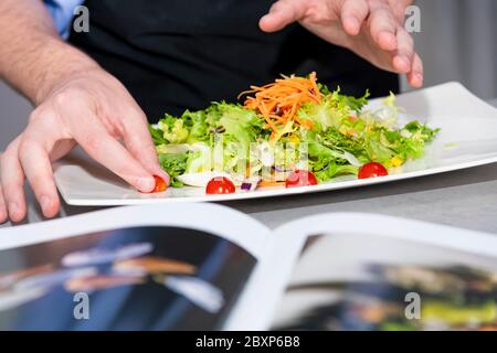 Nahaufnahme der männlichen Hände, die den letzten Schliff zu einem köstlich aussehenden Salat mit einem unscharf Buch an der Vorderseite. Kochen zu Hause und gesund f Stockfoto