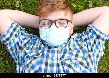 Porträt eines Jungen mit chirurgischer Maske auf dem Gras im Park liegend. Nahaufnahme eines kleinen Jungen im karierten Hemd, der sich auf dem Gras entspannt. Stockfoto