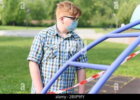 Junge mit OP-Maske vor einem rot-weiß gestreiften Sperrband um den Spielplatz. Fechten rot-weißes Band, Warnband. Stockfoto