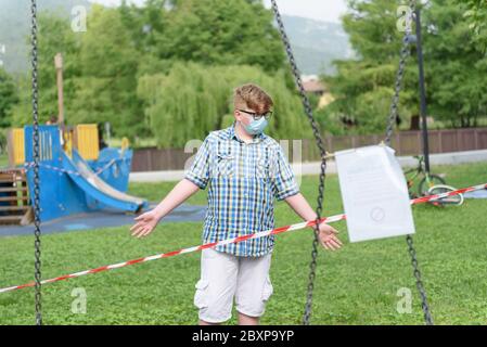 Junge mit OP-Maske vor einem rot-weiß gestreiften Sperrband um den Spielplatz. Fechten rot-weißes Band, Warnband. Stockfoto