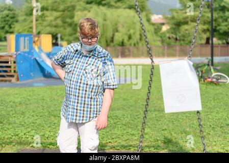 Junge mit OP-Maske vor einem rot-weiß gestreiften Sperrband um den Spielplatz. Fechten rot-weißes Band, Warnband. Stockfoto