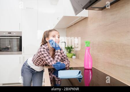 Ein müder junger Reiniger ruht nach dem Reinigen des Hauses in der Küche auf der Küchenarbeitspfläche.EIN junges Mädchen steht nachdenklich im Kittchen