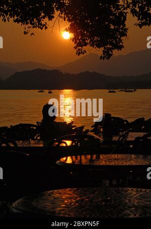 Sonnenuntergang am Westsee (Xi Hu) in Hangzhou, Provinz Zhejiang, China. Sonnenuntergang Blick auf West Lake, Hangzhou mit Silhouette einer Person in einem Café sitzen. Stockfoto