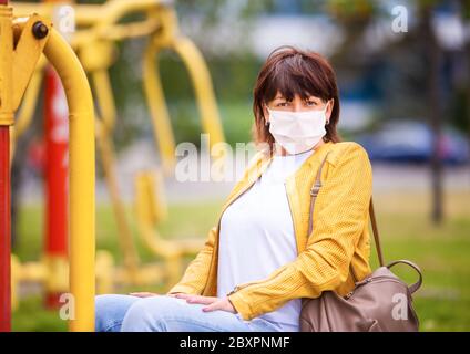 Unbekannte Frau mittleren Alters in legerer Kleidung geht in einer weißen Schutzmaske durch den Frühlingspark. Coronovirus-Schutzkonzept Stockfoto