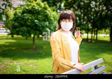 Frau mittleren Alters in einer schützenden weißen Maske geht im Park an einem sonnigen Frühlingstag warm. Coronovirus- und Quarantäneschutzkonzept Stockfoto