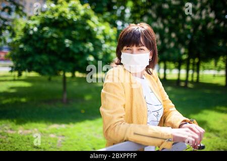 Frau mittleren Alters in einer schützenden weißen Maske geht im Park an einem sonnigen Frühlingstag warm. Coronovirus- und Quarantäneschutzkonzept Stockfoto