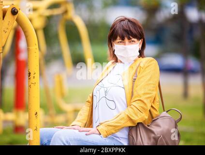 Unbekannte Frau mittleren Alters in legerer Kleidung geht in einer weißen Schutzmaske durch den Frühlingspark. Coronovirus-Schutzkonzept Stockfoto