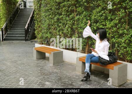Frau mit Gesichtsmaske auf einer Bank in einem Garten sitzend winkt Hand zu Freund Stockfoto