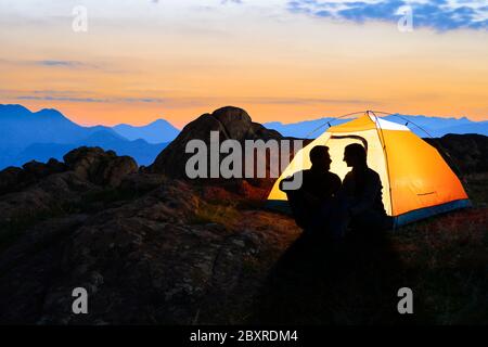 Junges Paar, das in der Nähe des beleuchteten Zeltes sitzt und sich am Beautirul Abend in den Bergen anschaut. Abenteuer und Familienreisen Konzept. Stockfoto