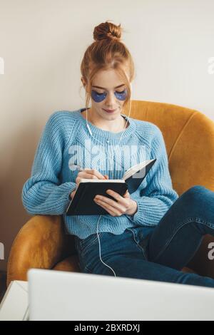 Rothaarige Dame mit Sommersprossen, die Hydrogel-Augenwege tragen, während sie Musik hört und ein Buch auf dem Sessel liest Stockfoto