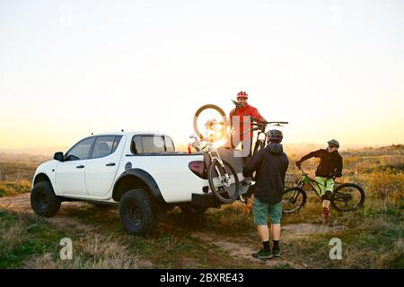 Freunde Radfahrer sich auf das Fahrradfahren vorbereiten und die Fahrräder vom Pickup Offroad Truck in den Bergen bei warmem Herbstuntergang abheben. MTB-Advent Stockfoto