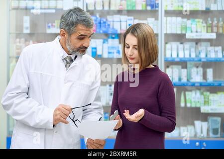 Vorderansicht der weiblichen Kundenberatung mit Chemiker, mit Blisterpackung von Pillen. Bärtiger Apotheker trägt Laborkittel, hilft jungen Frau in der Apotheke mit Medikamenten, erklärt Anweisung. Stockfoto