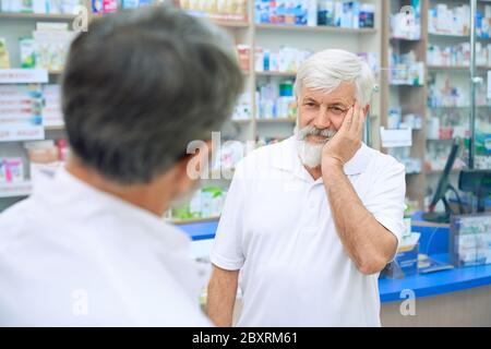 Selektive Fokussierung auf Senior man mit Zahnschmerzen Beratung mit Apotheker. Rückansicht des unkenntlichen Mannes in weißer Uniform, der Medikamente anbietet, während der elderige männliche Kunde schmerzhaft Stirnrunzeln. Stockfoto