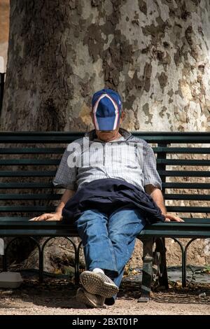 Ein Mann döst in der Sonne auf einer Parkbank mit seiner Mütze auf dem Gesicht. Stockfoto