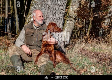 Alter Mann, der die Natur liebt, sitzt in der schönen Frühlingssonne draußen im Wald an einem Baum und hält seinen jungen irischen Setter in seinem Arm. Stockfoto