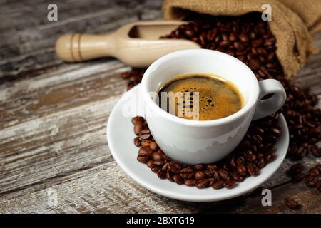 Kaffeetasse und geröstete Kaffeebohnen in einem Sackleinen auf einem rustikalen Holztisch Nahaufnahme auf rostigen Vintage-Hintergrund. Stockfoto