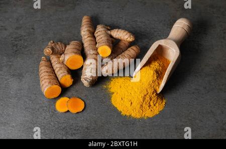 Kurkuma-Pulver gesunde Würze Asiatische Küche Nahaufnahme von Kurkuma Wurzel in Scheiben geschnitten und ein Holzbailer auf einem rustikalen dunkelgrauen Küchenbrett. Stockfoto