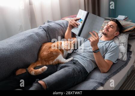 Weiße und braune Katze schläft auf seinem Besitzer ein, der auf einem Sofa unter dem Fenster liegend liest. Stockfoto