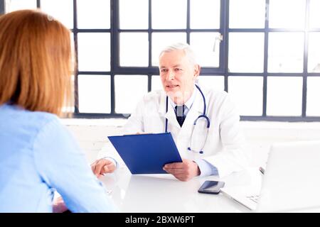 Schuss eines männlichen Arztes mittleren Alters, der mit seinem Patienten spricht, während er am Büroschreibtisch im Arztraum sitzt. Stockfoto