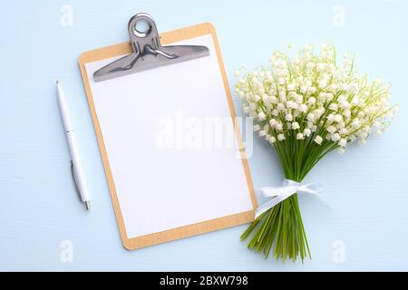 Clipboard mit leerem Papier Note, Stift, Blumenstrauß Lilie des Tals auf blauem Hintergrund. Flach liegend, Draufsicht. Hochzeitsplaner, Liste zu tun, zu überprüfen Stockfoto