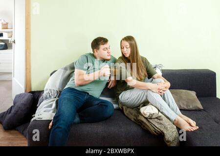 Junges Paar in lässiger Kleidung sitzen auf der Couch im Wohnzimmer Kaffee trinken und reden, bleiben Sie zu Hause Konzept, ganzen Körper Stockfoto