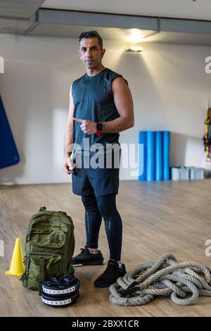 Boot Camp Instructor steht mit Fitnessgeräten in der Turnhalle. Hanteln, Wasserkocher, Seil, Sandsack auf Holzboden. Porträt des libanesischen Ausbilder für Stockfoto
