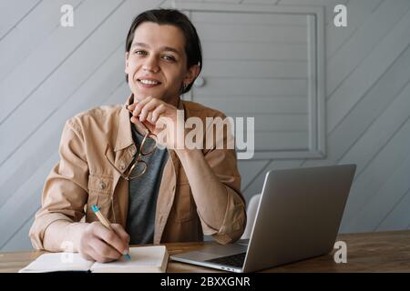 Lächelnder Mann schreibt Notizen in Notizbuch, arbeitet freiberuflich Projekt in Café. Junge Schüler mit Laptop, Studium, Prüfungsvorbereitung, Fernunterricht Stockfoto