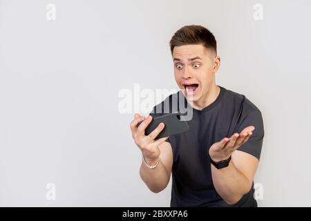 Ein überraschender, schockierter junger Mann hält ein Smartphone in den Händen und blickt mit offenem Mund und großen Augen auf das Display. Ein Kerl mit einem schlaffen Kiefer in Angst kam zuerst im Spiel, wurde ein Champion Stockfoto