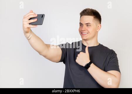 Porträt eines stilvollen gut aussehenden lächelnden jungen Mannes, der ein Selfie auf einem Smartphone, hält ein Handy in seiner ausgestreckten Hand und versucht, schön in Foto weißen Hintergrund Online-Konzept Stockfoto