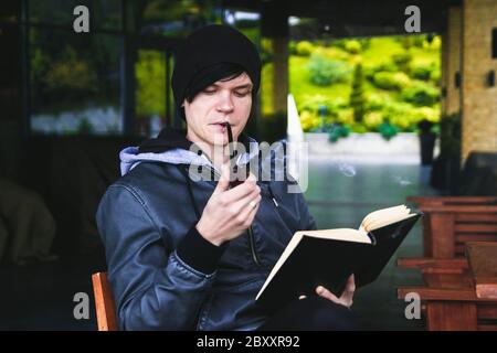 Junger schwarzhaariger Mann mit Piercing, der ein Buch reimt und Tabakpfeife im Café im Freien raucht Stockfoto