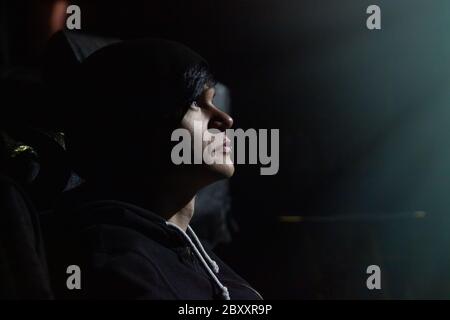 Die Seitenansicht des jungen schwarzhaarigen Mannes mit Augenbrauendurchbruch in der dunklen Halle des Konzerts oder Kinos Gesicht durch Bildschirm oder Szene Beleuchtung beleuchtet. Stockfoto